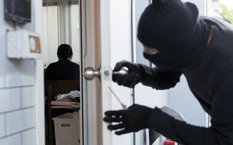 A masked intruder attempts to pick a lock on a door with a screwdriver